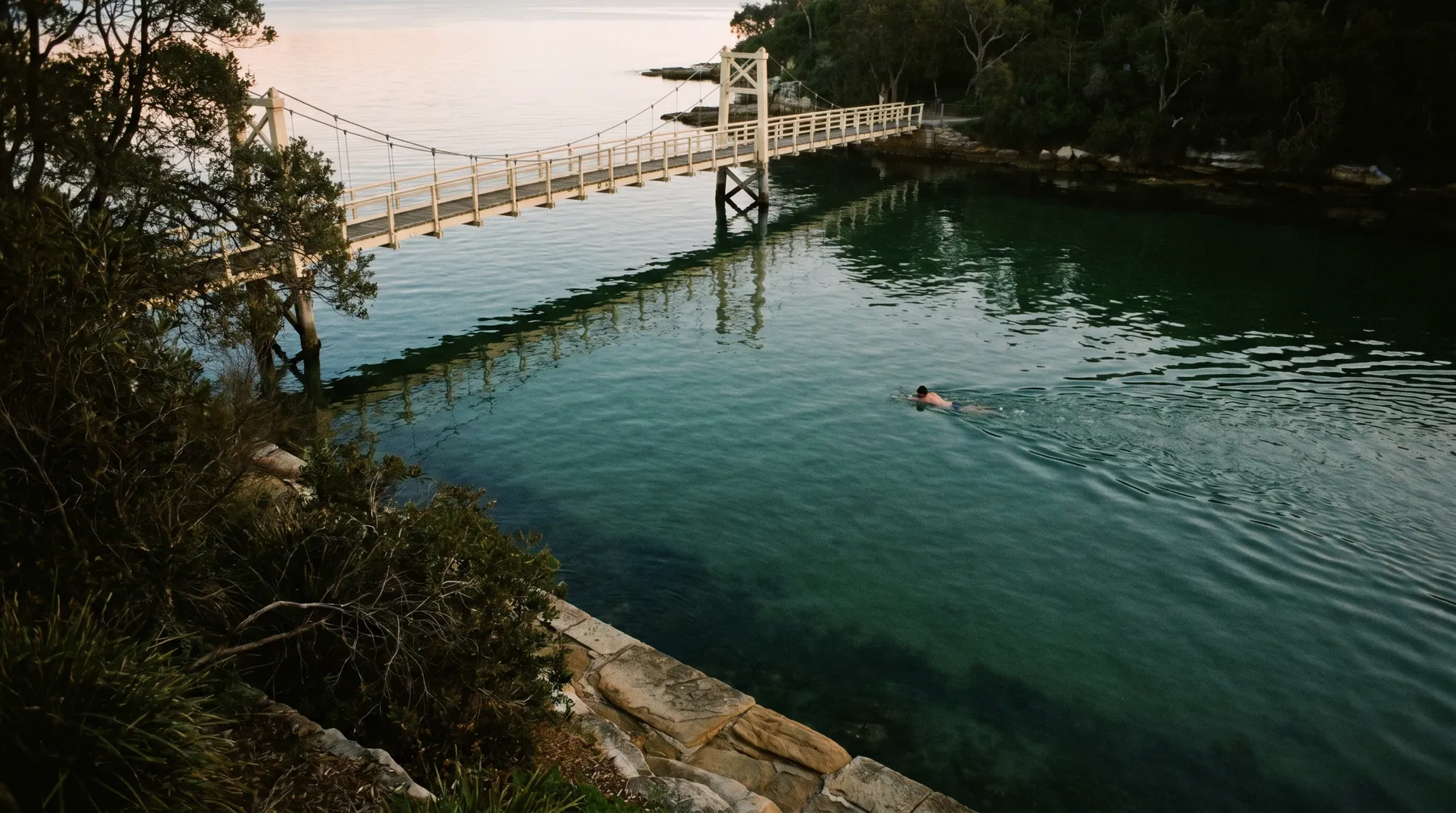 Parsley Bay's calm water and white suspension footbridge in early morning light
